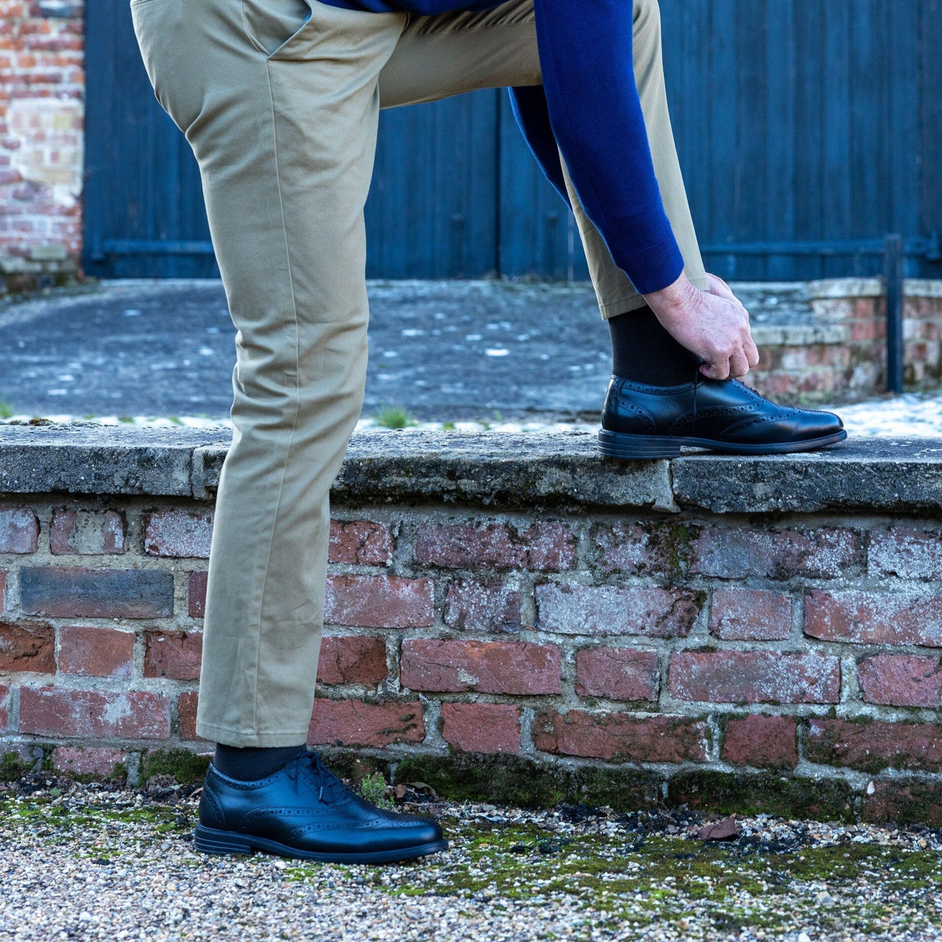 Man in a blue sweater and beige pants tying his shoe outdoors.